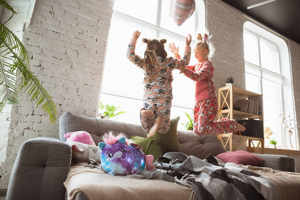 Quiet little girls playing in a bedroom in cute pajamas, home style and comfort, laughting and fighting pillows, jumping high - Photo, image