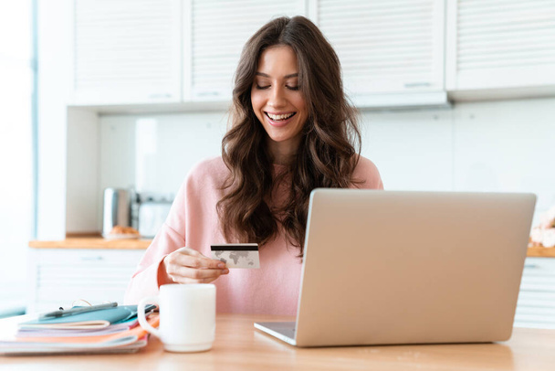 Image of a smiling cheerful beautiful young woman sit indoors at home using laptop computer holding credit card. - Foto, Imagem
