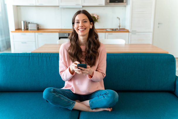 幸せな楽観的な若い女性のイメージはソファの上に携帯電話を使用して自宅で屋内に座る. - 写真・画像