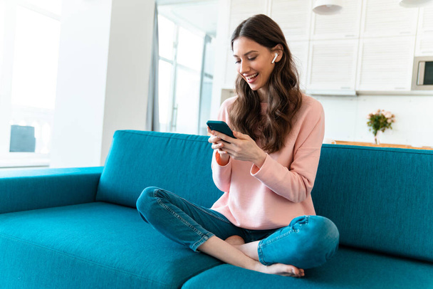Image of a happy optimistic young woman sit indoors at home using mobile phone on sofa. - Fotoğraf, Görsel