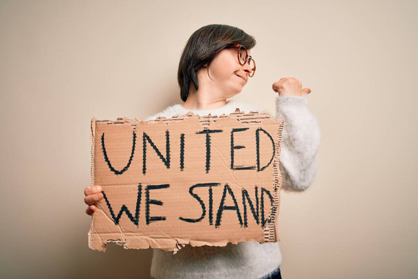 Young down syndrome woman holding protest banner with united we stand rights message pointing and showing with thumb up to the side with happy face smiling - Foto, imagen