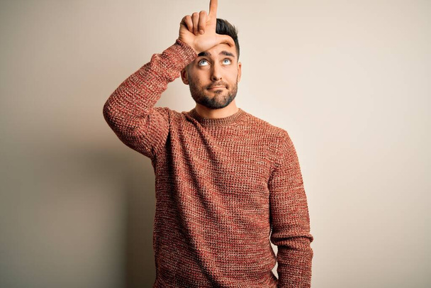 Young handsome man wearing casual sweater standing over isolated white background making fun of people with fingers on forehead doing loser gesture mocking and insulting. - Photo, Image