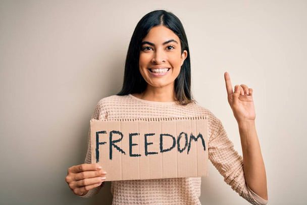 Young beautiful hispanic woman holding protest banner of freedom asking for liberty surprised with an idea or question pointing finger with happy face, number one - Foto, immagini