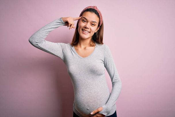Young beautiful teenager girl pregnant expecting baby over isolated pink background Smiling pointing to head with one finger, great idea or thought, good memory - 写真・画像