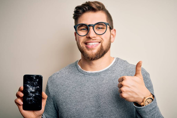 Mladý blonďatý muž s vousy a modrýma očima nosí brýle drží rozbitý smartphone šťastný s velkým úsměvem dělá ok znamení, palec nahoru s prsty, vynikající znamení - Fotografie, Obrázek