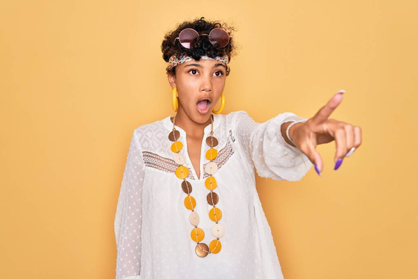 Young beautiful african american afro hippie woman wearing sunglasses and accessories Pointing with finger surprised ahead, open mouth amazed expression, something on the front - Foto, imagen