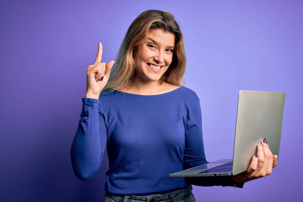 Fiatal gyönyörű szőke nő dolgozik laptoppal izolált lila háttér meglepett egy ötlet vagy kérdés ujjal mutogató boldog arc, első számú - Fotó, kép