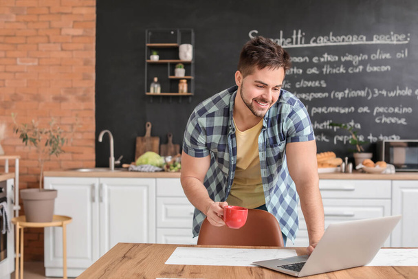 Young man with cup of hot coffee and laptop in kitchen - Fotoğraf, Görsel