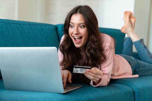 Image of a smiling emotional young woman indoors at home using laptop computer holding credit card on sofa. - Foto, Imagem