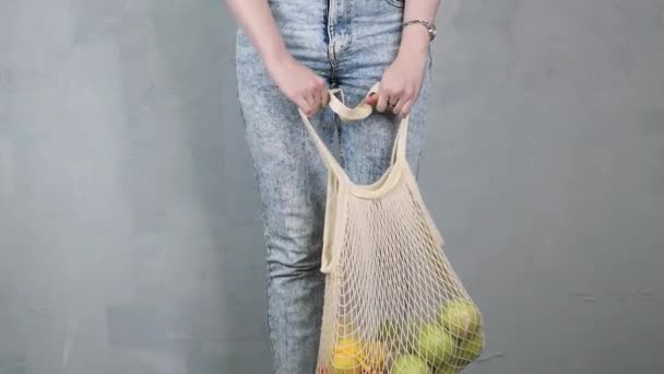 Nő kezében újrahasznosítható háló húr kötött bevásárló ökotáska gyümölcsökkel. Lábak kék farmerban. Szürke fal a háttérben - Felvétel, videó