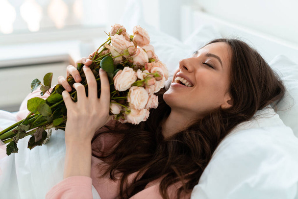 Image of a smiling pretty young woman indoors at home lies in bedroom in bed holding flowers. - Photo, Image