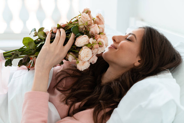 Image of a happy positive pretty young woman indoors at home lies in bedroom in bed holding flowers. - Fotografie, Obrázek