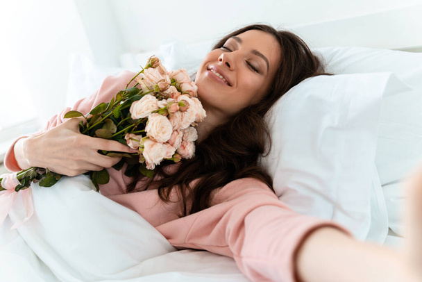 Afbeelding van een tevreden positieve mooie jonge vrouw binnen thuis ligt in de slaapkamer in bed met bloemen nemen een selfie met de camera. - Foto, afbeelding
