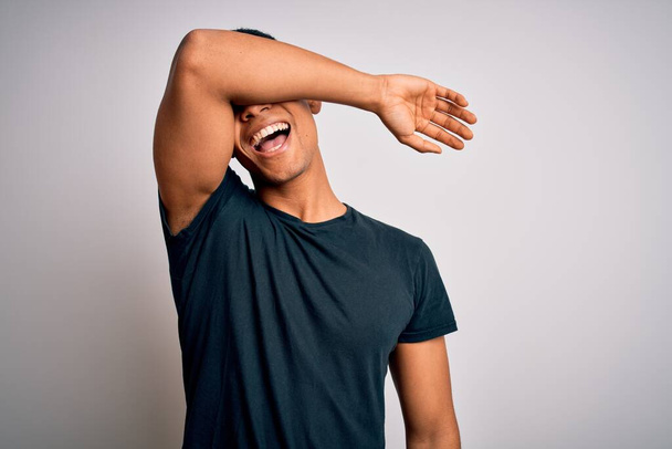 Joven hombre afroamericano guapo vistiendo camiseta casual de pie sobre fondo blanco cubriendo los ojos con el brazo sonriendo alegre y divertido. Concepto ciego
. - Foto, imagen