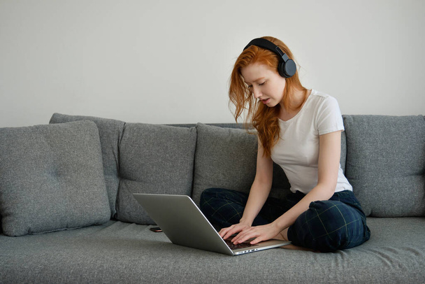 Redhead girl sits half-turned on the couch, legs crossed in home clothes and headphones, bowing head over the laptop, writes - Фото, изображение
