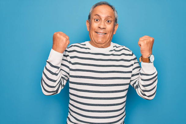 Senior handsome grey-haired man wearing striped sweater over isolated blue background celebrating surprised and amazed for success with arms raised and open eyes. Winner concept. - Photo, Image