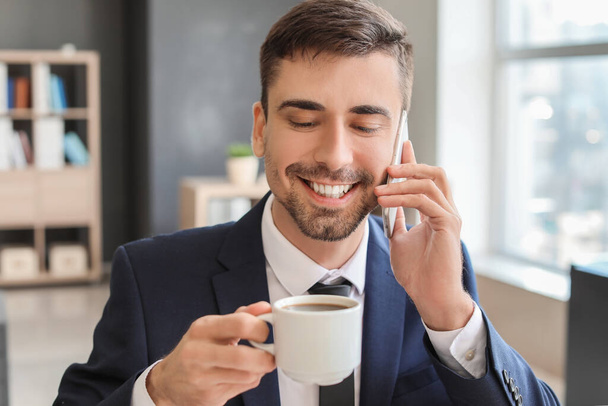 Młody biznesmen z filiżanką gorącej kawy rozmawia przez telefon w biurze - Zdjęcie, obraz