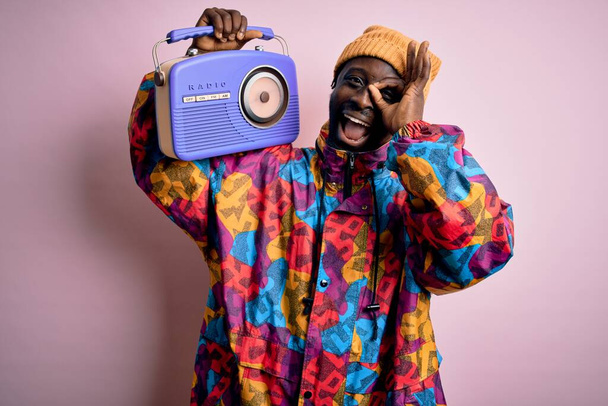 Jeune homme afro-américain écoutant de la musique tenant portable vintage radio rétro avec le visage heureux souriant faire ok signe avec la main sur les yeux regardant à travers les doigts
 - Photo, image