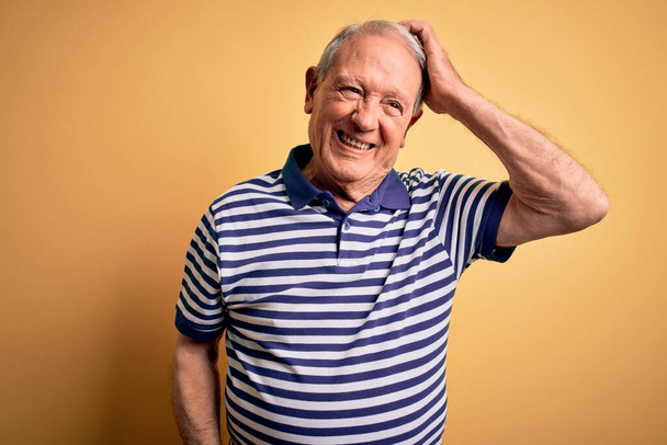 Grey haired senior man wearing casual navy striped t-shirt standing over yellow background confuse and wonder about question. Uncertain with doubt, thinking with hand on head. Pensive concept. - Foto, Imagen