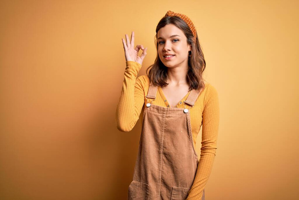 Young beautiful blonde girl wearing overall standing over yellow isolated background smiling positive doing ok sign with hand and fingers. Successful expression. - Photo, Image
