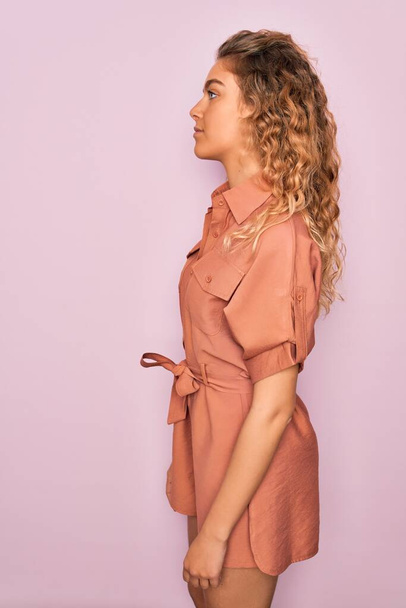 Jonge mooie vrouw met blauwe ogen dragen casual zomer jurk over roze achtergrond op zoek naar kant, ontspannen profiel pose met natuurlijke gezicht met zelfverzekerde glimlach. - Foto, afbeelding