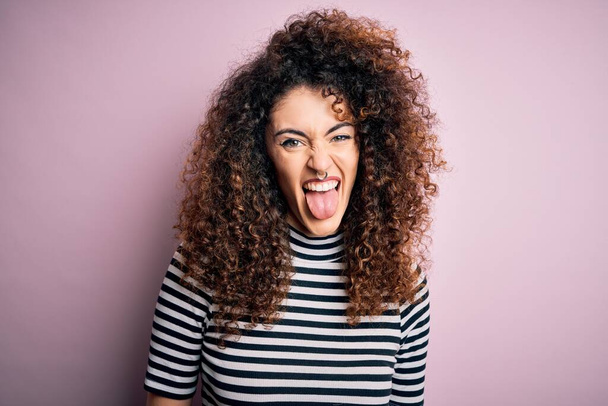 Молодая красивая женщина с вьющимися волосами и пирсинг носить случайные полосатые футболки торчащие язык счастливым от смешного выражения. Концепция эмоций
. - Фото, изображение