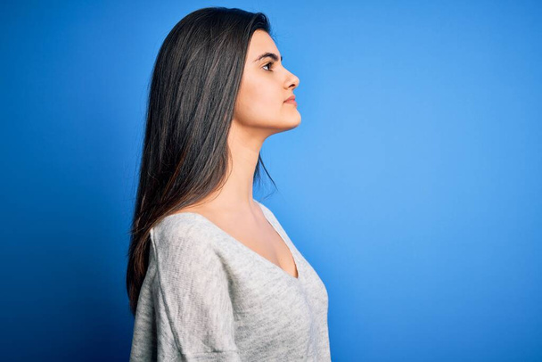 Молодая красивая брюнетка женщина в случайном свитере стоя на синем фоне глядя в сторону, расслабиться профиль позировать с естественным лицом с уверенной улыбкой
. - Фото, изображение