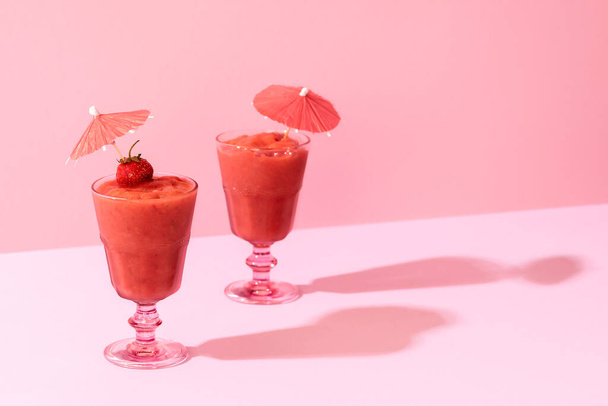 Дві полуничні смузі в склянці з коктейльною парасолькою на рожевому фоні, в суворому світлі. Літні смачні маки. Здоровий дієтичний напій
. - Фото, зображення