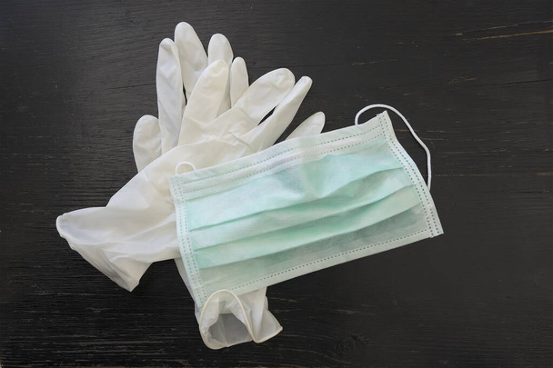 Ιατρική προστατευτική μάσκα και λευκά γάντια λατέξ πάνω από μαύρο ξύλινο φόντο. - Φωτογραφία, εικόνα