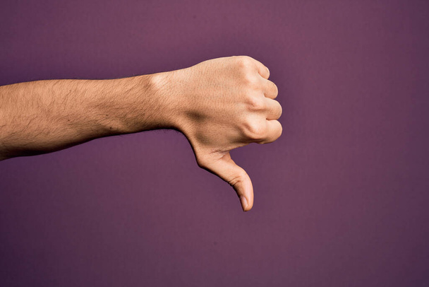 Mão de jovem caucasiano mostrando dedos sobre fundo roxo isolado fazendo polegares para baixo gesto de rejeição, desaprovação antipatia e sinal negativo - Foto, Imagem