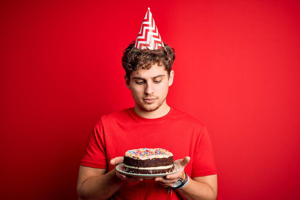 Νέος ξανθός άνδρας με σγουρά μαλλιά φορώντας καπέλο γενεθλίων κρατώντας τούρτα σε κόκκινο φόντο με μια σίγουρη έκφραση στο έξυπνο πρόσωπο σκέφτεται σοβαρά - Φωτογραφία, εικόνα