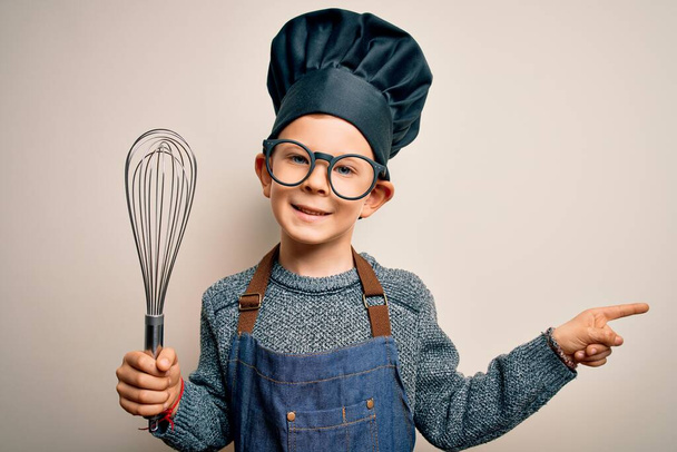Giovane bambino cuoco caucasico indossare uniforme chef e cappello con frusta manuale molto felice puntando con mano e dito di lato
 - Foto, immagini