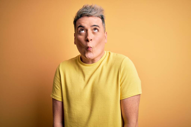 Young handsome modern man wearing yellow shirt over yellow isolated background making fish face with lips, crazy and comical gesture. Funny expression. - Fotó, kép