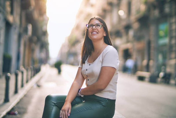 Mutlu ve kendinden emin gülümseyen genç ve güzel bir kadın. Kasaba caddesinde yüzünde gülümsemeyle dikilirken - Fotoğraf, Görsel