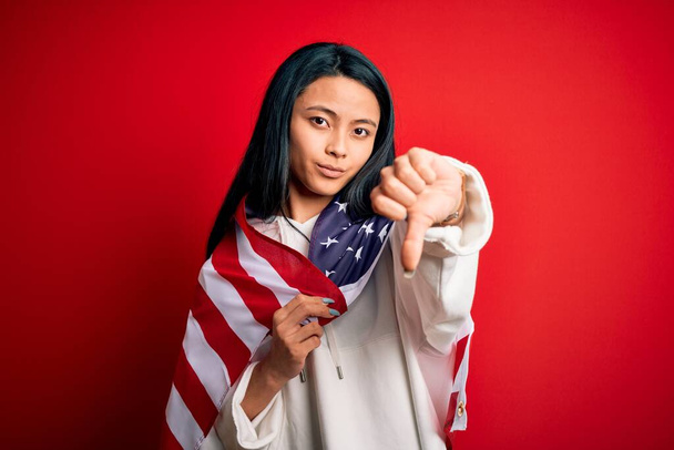 Young beautiful chinese woman wearing United States flag over isolated red background with angry face, negative sign showing dislike with thumbs down, rejection concept - Φωτογραφία, εικόνα