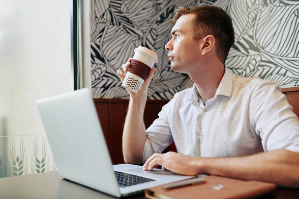 Молодий бізнесмен з чашкою п'є каву і дивиться через вікно після перевірки електронних листів на ноутбуці
 - Фото, зображення