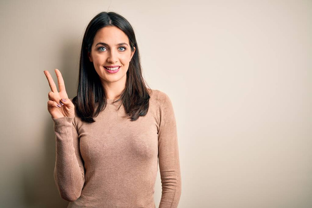 Young brunette woman with blue eyes wearing casual sweater over isolated white background smiling looking to the camera showing fingers doing victory sign. Number two. - Foto, Imagen