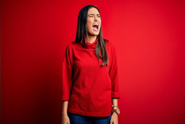 Young beautiful brunette woman wearing casual sweater over isolated red background angry and mad screaming frustrated and furious, shouting with anger. Rage and aggressive concept. - Photo, Image