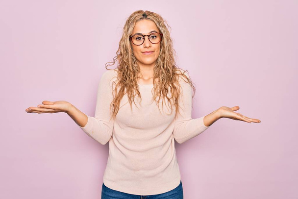 Young beautiful blonde woman wearing casual sweater and glasses over pink background clueless and confused expression with arms and hands raised. Doubt concept. - Photo, Image