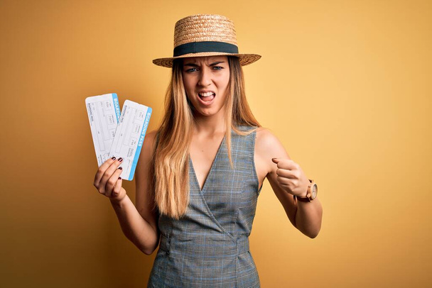 Young blonde tourist woman with blue eyes on vacation wearing hat holding boarding pass annoyed and frustrated shouting with anger, crazy and yelling with raised hand, anger concept - 写真・画像