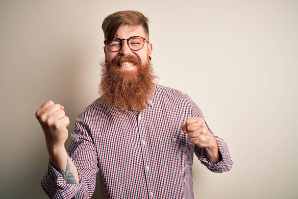 Komea irlantilainen punapää liikemies, jolla on parta silmälasit eristetyn taustan päällä, erittäin iloinen ja innoissaan tehdessään voittajan elettä kädet ylhäällä, hymyillen ja huutaen menestystä. Juhlan käsite
. - Valokuva, kuva