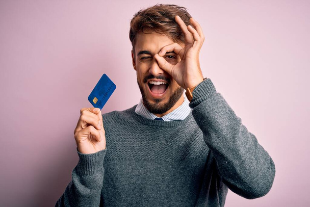 Fiatal ügyfél férfi szakáll tartja hitelkártya fizetési rózsaszín háttér boldog arc mosolygós csinál oké jel kéz a szemen keresztül néz az ujjak - Fotó, kép
