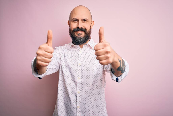 Schöner, glatzköpfiger Mann mit Bart, elegantem Hemd vor isoliertem rosafarbenem Hintergrund, der zustimmend positive Gesten mit der Hand macht, lächelnd Daumen hoch und glücklich über den Erfolg. Siegergeste. - Foto, Bild