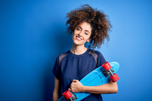 Jovem mulher esportiva com cabelo encaracolado e piercing segurando patins sobre fundo azul com um rosto feliz de pé e sorrindo com um sorriso confiante mostrando dentes
 - Foto, Imagem