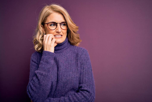 Middle age beautiful blonde woman wearing casual purple turtleneck sweater and glasses looking stressed and nervous with hands on mouth biting nails. Anxiety problem. - Photo, Image