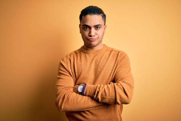 Young brazilian man wearing casual sweater standing over isolated yellow background skeptic and nervous, disapproving expression on face with crossed arms. Negative person. - Photo, Image