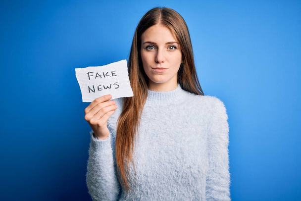 Young beautiful redhead woman holding paper with fake news message over blue background with a confident expression on smart face thinking serious - Fotó, kép