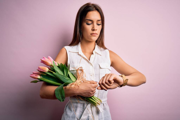 Joven mujer morena hermosa sosteniendo ramo de tulipanes flores sobre fondo rosa Comprobación de la hora en reloj de pulsera, relajado y seguro
 - Foto, Imagen