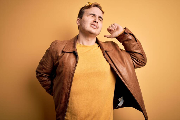 Young handsome redhead man wearing casual leather jacket over isolated yellow background Suffering of neck ache injury, touching neck with hand, muscular pain - Photo, image