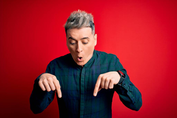 Mladý pohledný moderní muž v elegantní zelené košili přes červené izolované pozadí ukazuje dolů s prsty ukazující reklamu, překvapený obličej a otevřená ústa - Fotografie, Obrázek